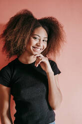 Lächelnde Afro-Frau mit Hand am Kinn vor einem pfirsichfarbenen Hintergrund - EBBF02504
