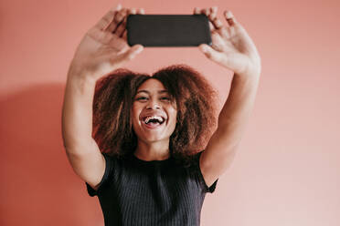 Fröhliche Afro-Frau nimmt Selfie durch Handy gegen Pfirsich Hintergrund - EBBF02506