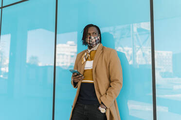 African man in protective face mask with smart phone standing while leaning on blue glass wall - EGAF01791