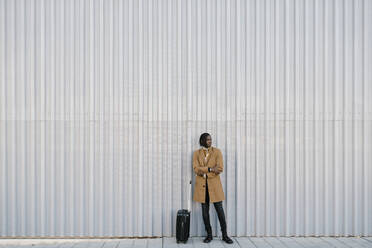 Man with suitcase standing against gray wall while looking away - EGAF01795