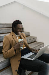 Glücklicher Mann mit Kaffeetasse und Laptop auf einer Treppe sitzend - EGAF01809