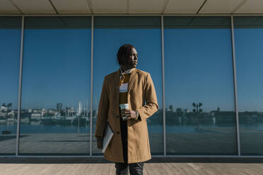 Afrikanischer Mann mit Laptop, der an einer Glaswand steht und wegschaut - EGAF01814