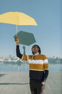 Lächelnder Mann mit Hand in der Tasche, der einen gelben Regenschirm hält und an einer Glaswand steht - EGAF01818