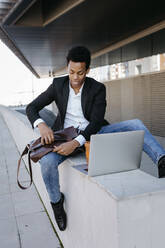 Geschäftsmann mit Laptoptasche auf Stützmauer sitzend - TCEF01544