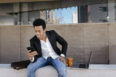 Mittlerer erwachsener Geschäftsmann, der sein Smartphone benutzt, während er auf einer Stützmauer sitzt - TCEF01554