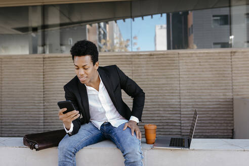 Mid adult businessman using smart phone while sitting on retaining wall - TCEF01554