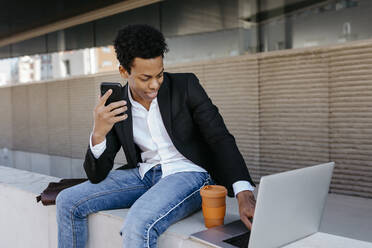 Lächelnder männlicher Unternehmer mit Smartphone und Laptop, der auf einer Stützmauer sitzt - TCEF01556