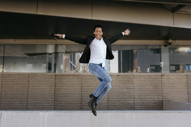 Cheerful male entrepreneur with arms outstretched jumping from retaining wall - TCEF01562