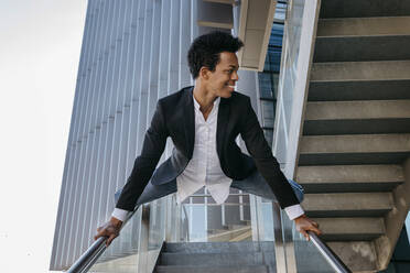 Smiling flexible businessman doing splits on staircase railing - TCEF01565