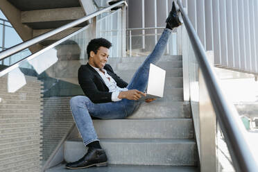 Flexibler mittelgroßer Geschäftsmann, der sein Bein ausstreckt und einen Laptop benutzt, während er auf einer Treppe sitzt - TCEF01568