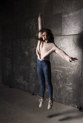 Happy young girl jumping with hand raised against wall - DAMF00722