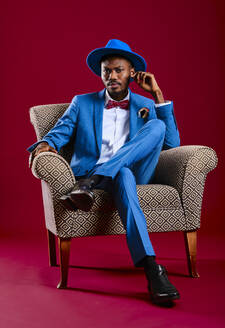 Confident man wearing blue suit sitting on chair against red background - AGOF00057