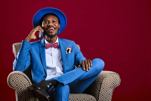 Laughing man talking on smart phone while sitting on chair against red background - AGOF00058