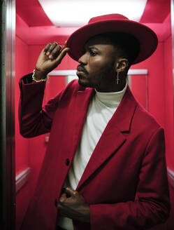 African fashionable man holding hat while peeking outside elevator - AGOF00064