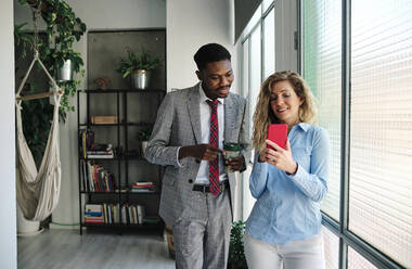 Businessman with coffee cup looking at smart phone held by female colleague while standing by glass window - AGOF00068