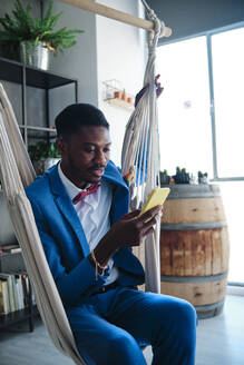 Mid adult man using smart phone while sitting on hammock - AGOF00071