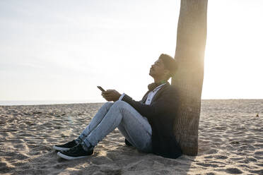 Entspannter Mann mit Smartphone, der an einem sonnigen Tag an einem Baum am Strand sitzt - JRFF05038