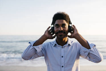 Lächelnder Afro-Mann hört Musik über Kopfhörer am Strand gegen den klaren Himmel - JRFF05051