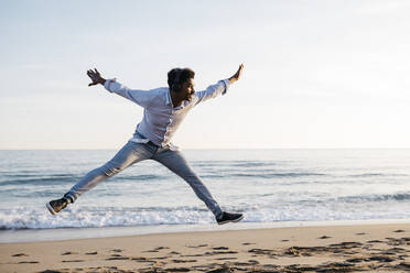 Cheerful man with arms outstretched jumping at beach against clear sky - JRFF05053