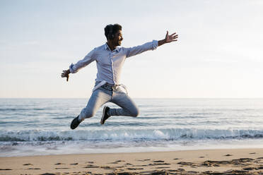 Happy man gesturing while jumping at beach against clear sky - JRFF05054