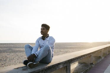 Thoughtful man sitting on retaining wall at beach against clear sky during sunny day - JRFF05058