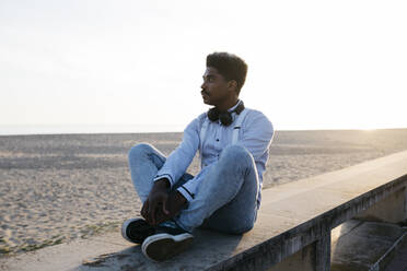 Contemplating man sitting on retaining wall at beach against clear sky during sunny day - JRFF05059