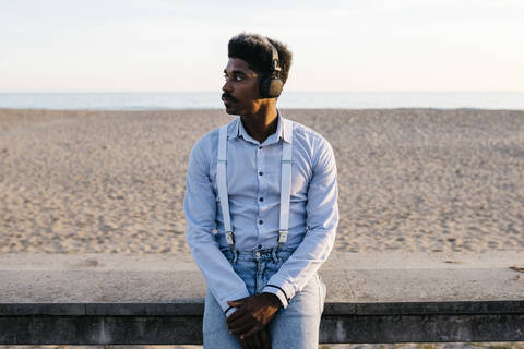 Gut aussehender Afro-Mann, der Musik über Kopfhörer hört, während er sich am Strand an eine Stützmauer lehnt, lizenzfreies Stockfoto