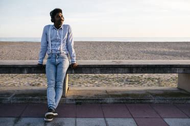 Smiling man with headphones leaning against retaining wall at beach - JRFF05062