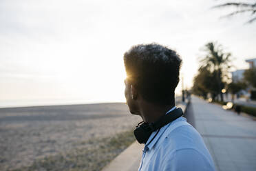 Afro-Mann mit Kopfhörern, der bei Sonnenuntergang die Aussicht vom Strand betrachtet - JRFF05064