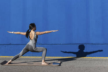 Woman doing Virabhadrasana pose in front of blue wall - PNAF00687