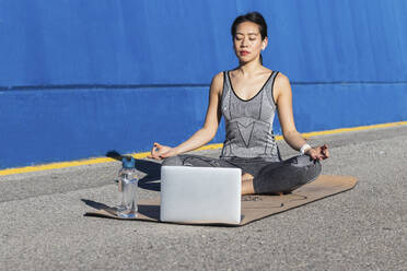 Frau im Lotussitz lernt Yoga, während sie vor einem Laptop sitzt - PNAF00690