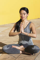 Beautiful woman with hands clasped meditating against yellow wall - PNAF00701