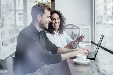 Lächelnde Geschäftsfrau mit Mobiltelefon neben einem Kollegen, der in einem Café am Laptop arbeitet - JOSEF03455