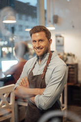 Owner wearing apron smiling while standing with arms crossed at cafe - JOSEF03470