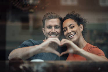 Smiling couple making heart with hand while sitting by cafe window - JOSEF03517