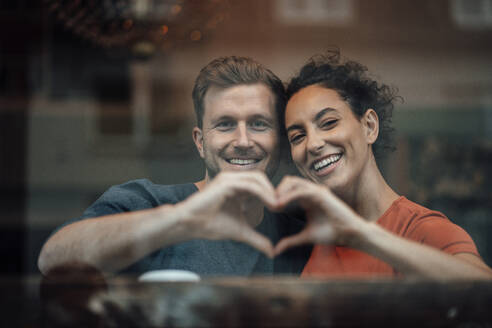 Lächelndes Paar macht ein Herz mit der Hand, während es am Fenster eines Cafés sitzt - JOSEF03517