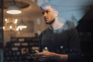 Mid adult man using mobile phone while standing by cafe window - JOSEF03522