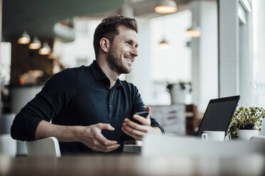 Lächelnder Geschäftsmann mit Laptop und Mobiltelefon, der in einem Café sitzt und wegschaut - JOSEF03527