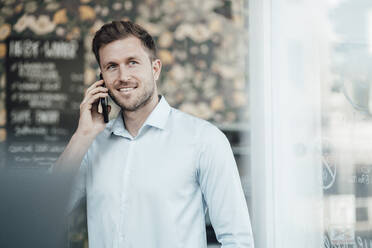 Lächelnder Geschäftsmann, der in einem Café steht und mit seinem Handy telefoniert - JOSEF03540