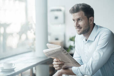 Smiling businessman with newspaper looking away while standing at cafe - JOSEF03543