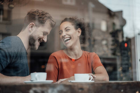 Freundin und Freund lachen, während sie am Fenster eines Cafés sitzen - JOSEF03562