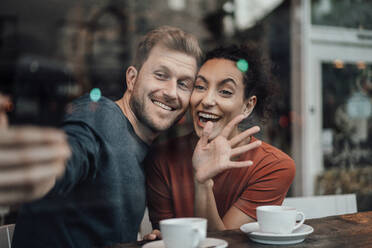 Lächelndes Paar, das einen Videoanruf auf dem Mobiltelefon betrachtet, während es im Café sitzt - JOSEF03565