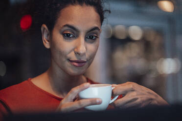 Junge Frau trinkt Kaffee, während sie in einem Café sitzt und auf ihren Laptop schaut - JOSEF03588