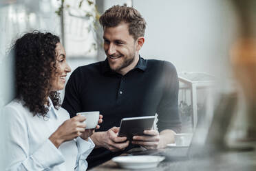 Lächelnder Geschäftsmann, der ein digitales Tablet benutzt, während er neben einem Kollegen steht, der in einem Café Kaffee trinkt - JOSEF03604
