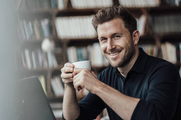 Lächelnder Geschäftsmann trinkt Kaffee und sitzt mit einem Laptop in einem Café - JOSEF03607