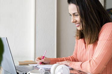 Smiling female entrepreneur writing in diary during video call at home office - AFVF08175