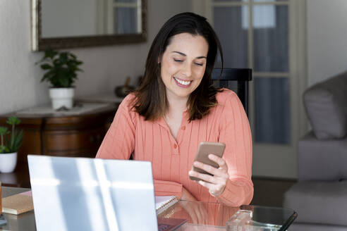 Lächelnde Geschäftsfrau, die auf ihr Handy schaut, während sie am Esstisch im Heimbüro sitzt - AFVF08177