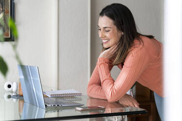 Happy female entrepreneur with head in hand leaning on dining table during video call at home - AFVF08180