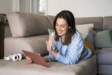 Glückliche Frau, die über ein digitales Tablet einen Videoanruf führt, während sie zu Hause auf dem Sofa liegt - AFVF08184