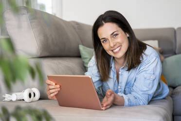 Schöne Frau lächelt, während sie ein digitales Tablet auf dem Sofa zu Hause hält - AFVF08185